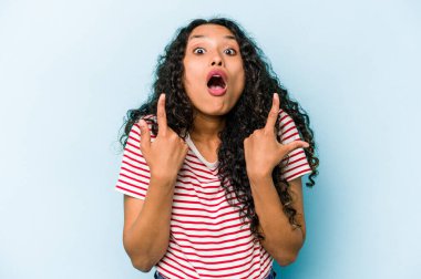 Mavi arka planda izole edilmiş genç İspanyol kadın ağzı açık bir şekilde üst tarafı işaret ediyor..