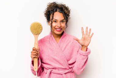 Sırt kaşıyıcı tutan genç İspanyol kadın beyaz arka planda izole edilmiş, gülümseyen beş numarayı parmaklarıyla gösteriyor..