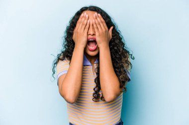 Mavi arka planda izole edilmiş genç İspanyol kadın yüzünün yarısını avuç içiyle kaplayarak eğleniyor..