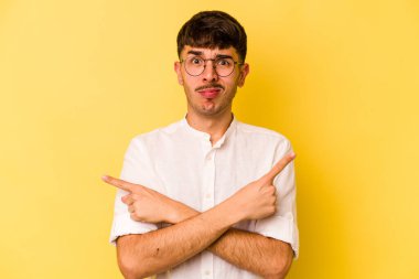 Sarı arka planda izole genç beyaz adam, iki seçenek arasında seçim yapmaya çalışıyor.