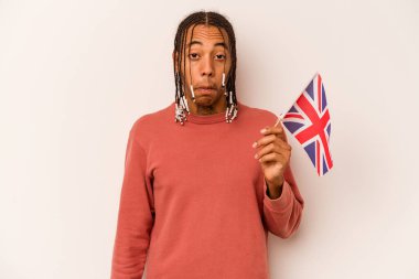 Beyaz arka planda izole edilmiş İngiliz bayrağı tutan genç Afrikalı Amerikalı omuz silkiyor ve gözleri açık..