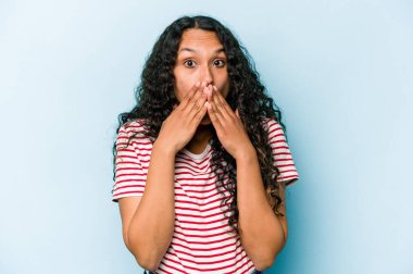 Mavi arka planda izole edilmiş genç İspanyol kadın elleriyle ağzını kapatırken şok oldu..
