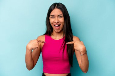 Mavi arka planda izole edilmiş genç İspanyol kadın parmağını göstererek şaşırdı, geniş bir gülümsemeyle..