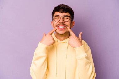 Mor arka planda izole edilmiş genç beyaz adam gülümsüyor, parmaklarını ağıza doğrultuyor..