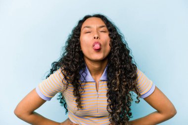 Mavi arka planda izole edilmiş genç bir İspanyol kadın. Komik ve dost canlısı bir dil uzatıyor..