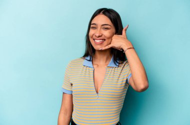 Mavi arka planda izole edilmiş genç İspanyol kadın parmaklarıyla bir cep telefonu hareketi gösteriyor..