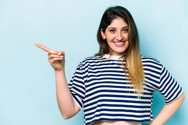 Mavi arka planda izole edilmiş genç beyaz kadın işaret parmağıyla işaret ederek gülümsüyor..