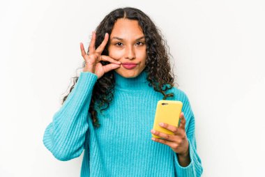 Cep telefonu tutan Latin kökenli genç bir kadın beyaz arka planda izole edilmiş ve parmakları dudaklarında bir sır saklıyor..