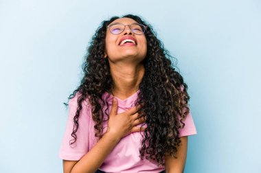 Mavi arka planda izole edilmiş genç İspanyol kadın yüksek sesle gülüyor elini göğsüne koyuyor..