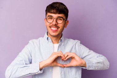Mor arka planda izole edilmiş genç beyaz adam gülümsüyor ve elleriyle kalp şeklini gösteriyor..