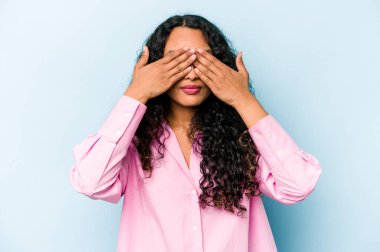 Mavi arka planda izole edilmiş genç İspanyol kadın elleriyle gözlerini kapatmaktan korkuyor..