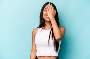 Mavi arka planda izole edilmiş genç İspanyol kadın yüzünün yarısını avuç içiyle kaplayarak eğleniyor..