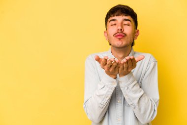 Sarı arka planda izole edilmiş genç, beyaz bir adam. Dudaklarını kıvırıyor, avuç içlerini tutuyor ve hava öpücüğü yolluyor..