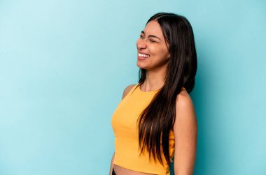 Mavi arka planda izole edilmiş genç İspanyol kadın gevşemiş ve mutlu bir şekilde gülüyor, boynunda dişlerini gösteriyor..