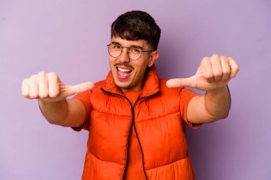 Mor arka planda izole edilmiş genç beyaz adam iki parmağını da kaldırıyor, gülümsüyor ve kendinden emin..