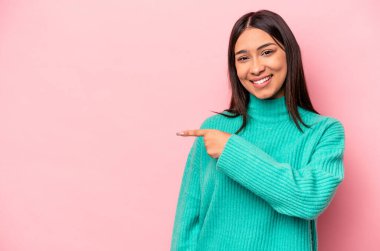 Pembe arka planda izole edilmiş genç İspanyol kadın gülümsüyor ve boş alanda bir şeyler gösteriyor..