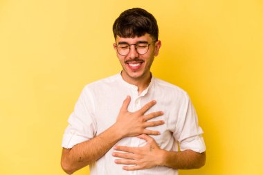 Sarı arka planda izole edilmiş genç beyaz adam mutlu bir şekilde gülüyor ve ellerini karnının üstünde tutarak eğleniyor..