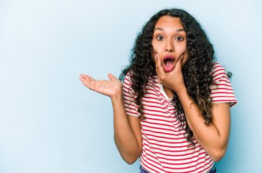 Mavi arka planda izole edilmiş genç İspanyol kadın avuç içinde fotokopi alanı tutuyor, el üstünde tutuyor. Şaşırmış ve mutlu..