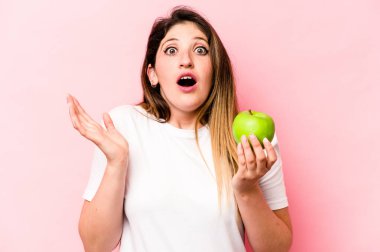 Elma tutan beyaz bir kadın pembe arka planda soyutlanmış, şaşırmış ve şok olmuş..