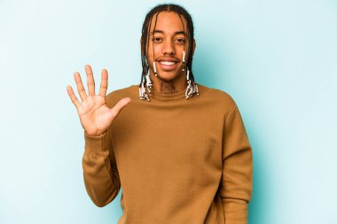 Genç Afro-Amerikalı adam mavi arka planda izole edilmiş. Gülümsüyor. Beş numarayı parmaklarıyla gösteriyor..