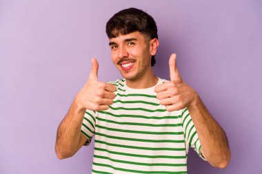 Mor arka planda izole edilmiş genç beyaz adam iki parmağını da kaldırıyor, gülümsüyor ve kendinden emin..