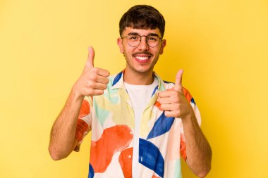 Sarı arka planda izole edilmiş genç beyaz adam iki parmağını da kaldırıyor, gülümsüyor ve kendinden emin..