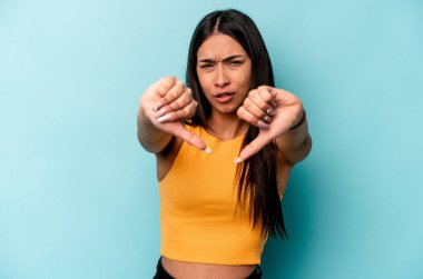 Mavi arka planda izole edilmiş genç İspanyol kadın baş parmağını aşağı gösteriyor ve hoşlanmadığını ifade ediyor..