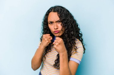 Mavi arka planda izole edilmiş genç İspanyol kadın kameraya yumruk gösteriyor, agresif yüz ifadesi..