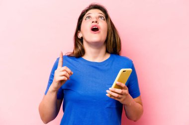 Elinde cep telefonu olan beyaz bir kadın pembe arka planda izole edilmiş ve ağzı açık bir şekilde üst tarafı işaret ediyor..