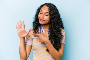Mavi arka planda izole edilmiş genç İspanyol kadın gülümseyerek beş numarayı parmaklarıyla gösteriyor..