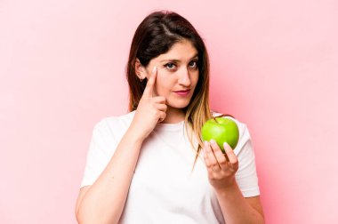 Elma tutan beyaz bir kadın pembe arka planda izole edilmiş, parmağıyla şakağı işaret ediyor, düşünüyor, bir göreve odaklanıyor..