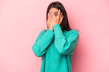 Pembe arka planda izole edilmiş genç İspanyol kadın parmaklarının arasından korkmuş ve gergin bir şekilde göz kırpıyor..