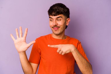 Mor arka planda izole edilmiş genç beyaz adam gülümseyerek beş numarayı parmaklarıyla gösteriyor..