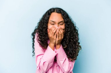 Mavi arka planda izole edilmiş genç İspanyol kadın bir şeye gülüyor, elleriyle ağzını kapatıyor..