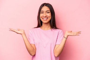 Pembe arka planda izole edilmiş genç İspanyol kadın kollarıyla ölçeği yapıyor, mutlu ve kendinden emin hissediyor..