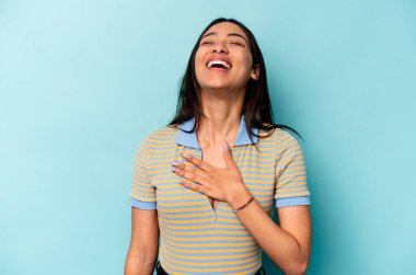 Mavi arka planda izole edilmiş genç İspanyol kadın yüksek sesle gülüyor elini göğsüne koyuyor..