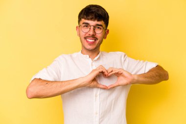 Sarı arka planda izole edilmiş genç beyaz adam gülümsüyor ve elleriyle kalp şeklini gösteriyor..