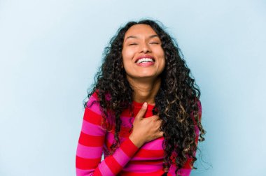 Mavi arka planda izole edilmiş genç İspanyol kadın yüksek sesle gülüyor elini göğsüne koyuyor..