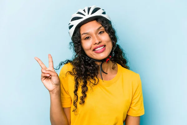 青い背景に隔離されたヘルメットバイクを身に着けている若いヒスパニック女性喜びと屈託のない指で平和のシンボルを示す — ストック写真