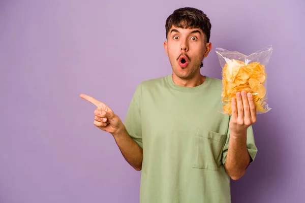 Jonge Spaanse Man Met Chips Geïsoleerd Paarse Achtergrond Wijst Naar — Stockfoto