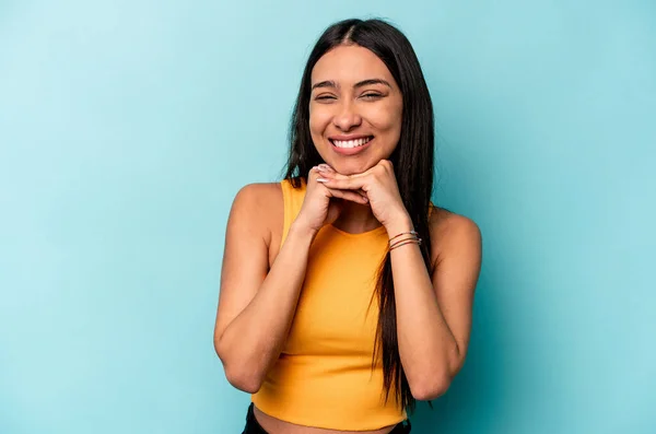 Young hispanic woman isolated on blue background keeps hands under chin, is looking happily aside.