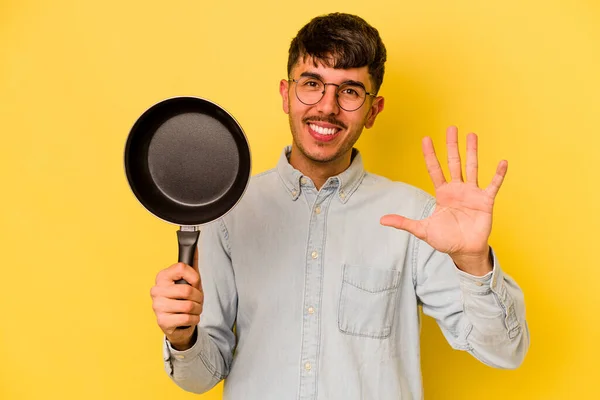 若いですヒスパニック炊飯器保持フライパン隔離上の黄色の背景笑顔陽気なショー番号5とともに指 — ストック写真