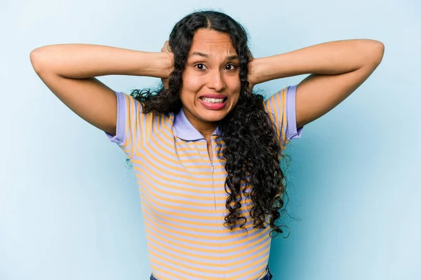 Young hispanic woman isolated on blue background covering ears with hands trying not to hear too loud sound.