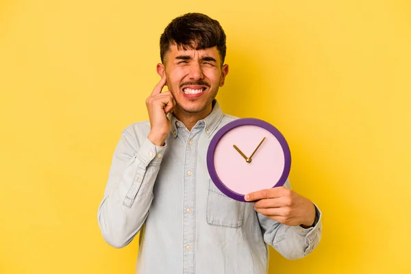 Молодий Іспаномовний Чоловік Тримає Годинник Ізольований Жовтому Тлі Покриваючи Вуха — стокове фото