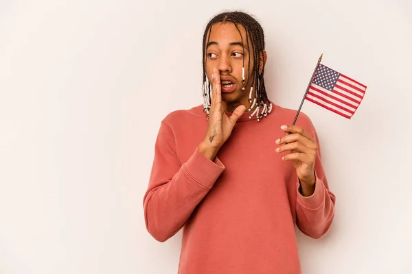 Young African American man holding a American flag isolated on white background is saying a secret hot braking news and looking aside