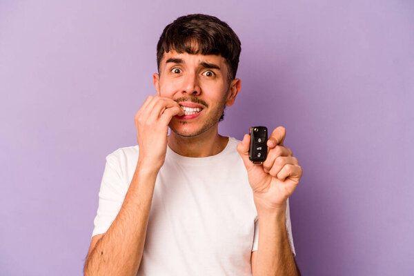 Young hispanic man holding keys car isolated on purple background biting fingernails, nervous and very anxious.