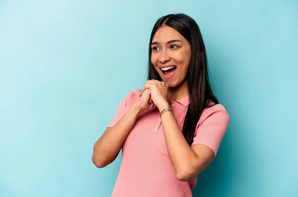 Young hispanic woman isolated on blue background keeps hands under chin, is looking happily aside.