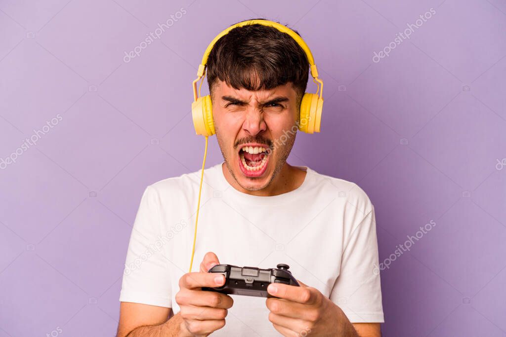 Young hispanic man playing with a video game controller isolated on purple background screaming very angry and aggressive.