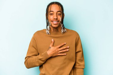 Mavi arka planda izole edilmiş genç Afro-Amerikalı adam yüksek sesle gülüyor ve elini göğsüne koyuyor..