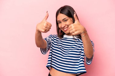 Genç İspanyol bir kadın pembe arka planda izole edilmiş. Baş parmağını kaldırıyor, gülümsüyor ve kendinden emin..